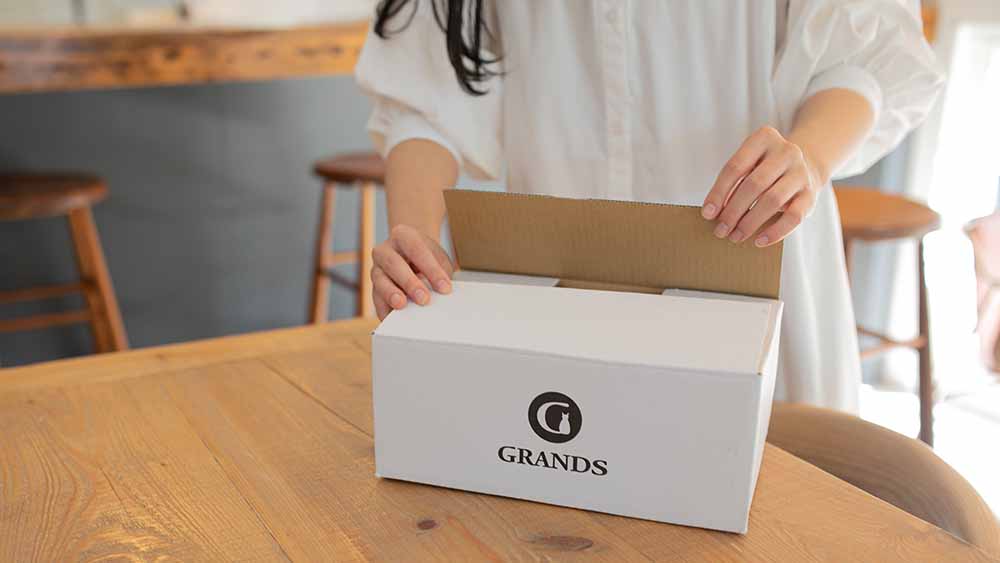 GRANDS（グランツ）のロゴが印字された専用のダンボール箱を開け、新鮮なドッグフードを受け取る飼い主の様子