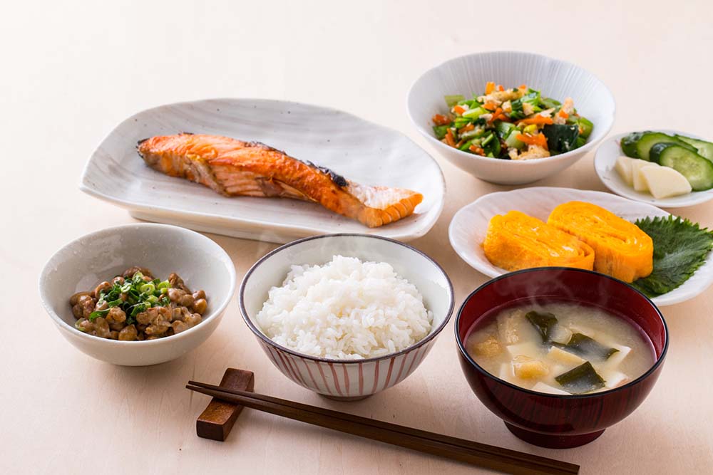 人間の食事の写真。ご飯・お味噌汁・納豆・卵焼き・焼き鮭・サラダ・きゅうりと大根のお漬物がある。