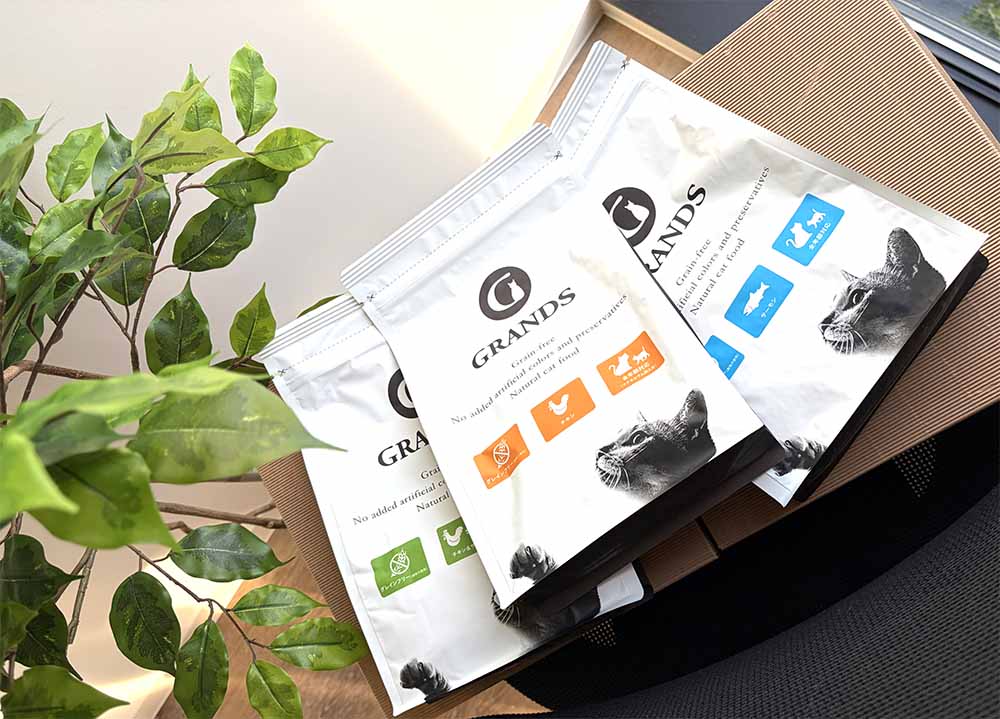 テーブルの上にグランツが販売しているキャットフード3種類（チキン&サーモン味とチキン味とサーモン味）のパッケージが横一列に並べられている写真。左には観葉植物が見切れている。