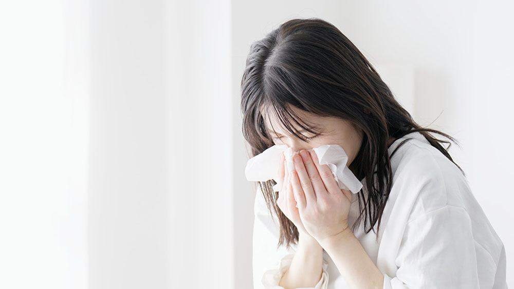 ティッシュで鼻をかむ女性。くしゃみ、鼻水、目のかゆみなど、アレルギーの代表的な症状を表現。
